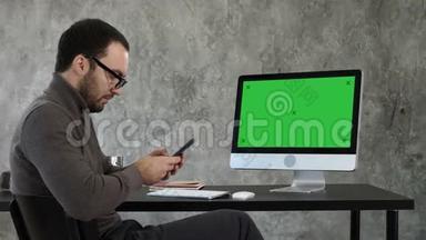 商人看着他的智能手机短信，坐在电脑屏幕附近。 绿色屏幕模拟显示。